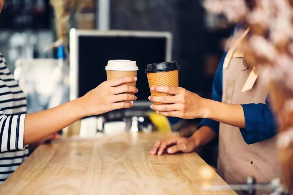 Barista handing a takeaway paper coffee cup to a customer across a wooden coffee shop counter.