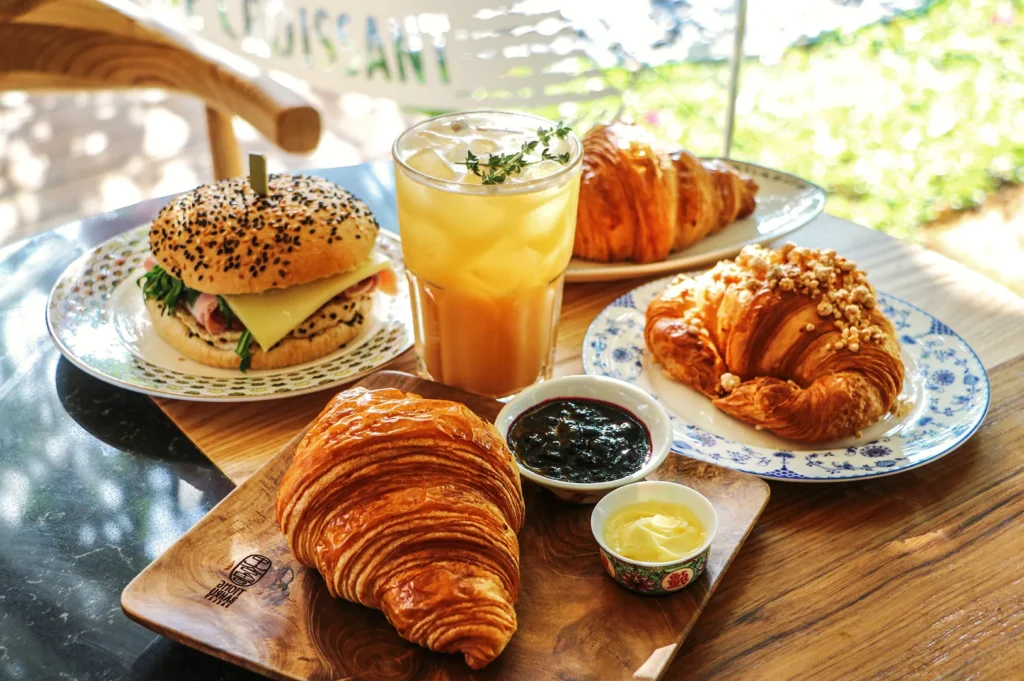 Breakfast spread with croissants, sandwich, jam, butter, and iced citrus drink — a bright café table showcasing fresh pastries and morning delights.