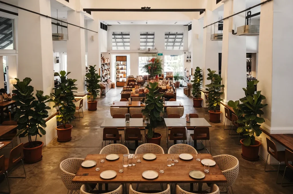 Spacious restaurant interior with high ceilings, wooden tables, and potted plants — a modern dining space combining natural light and minimalist design.