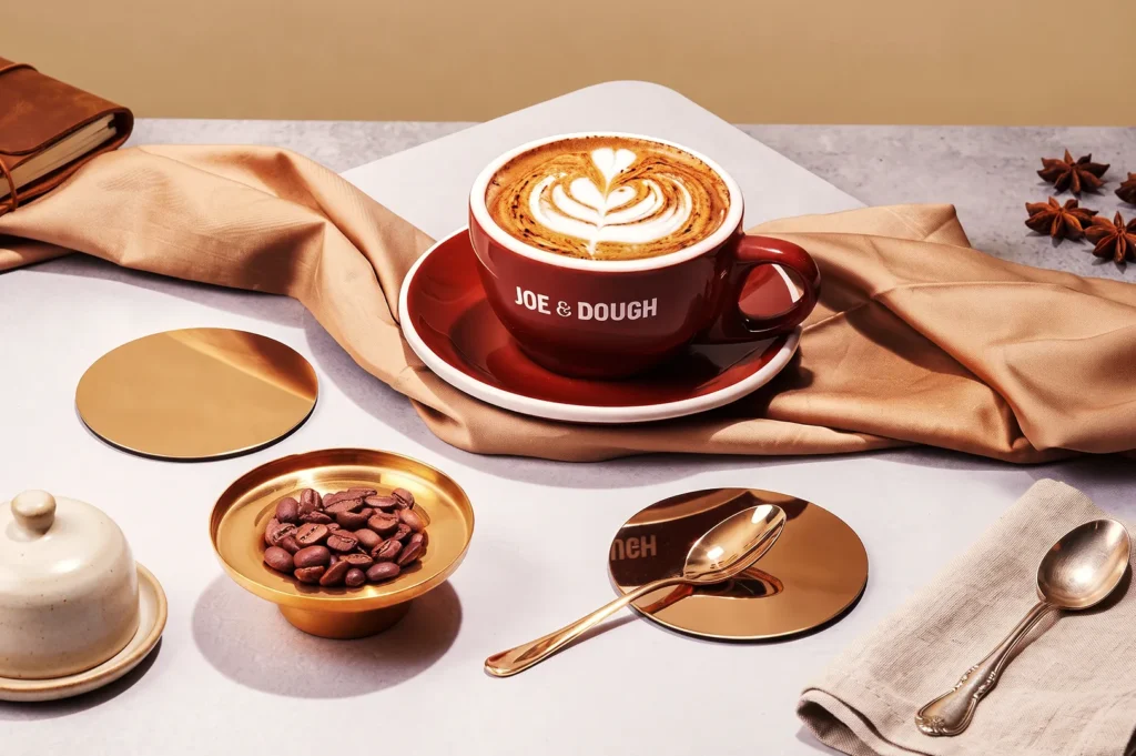Joe & Dough cappuccino with heart‑shaped latte art served in a red cup surrounded by golden tableware — premium handcrafted coffee presentation.