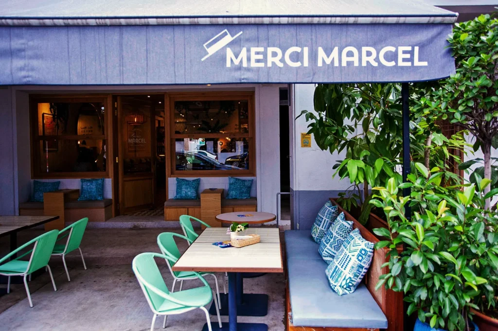 Outdoor seating area of Merci Marcel café with mint‑green chairs, patterned pillows, and lush greenery under a gray awning, creating a vibrant alfresco dining atmosphere.
