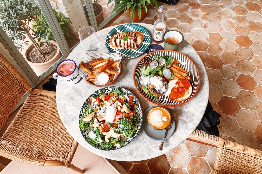 Vibrant brunch spread with salad, burger, fries, and coffee served on patterned plates in a sunlit café with terracotta floors and wicker chairs.