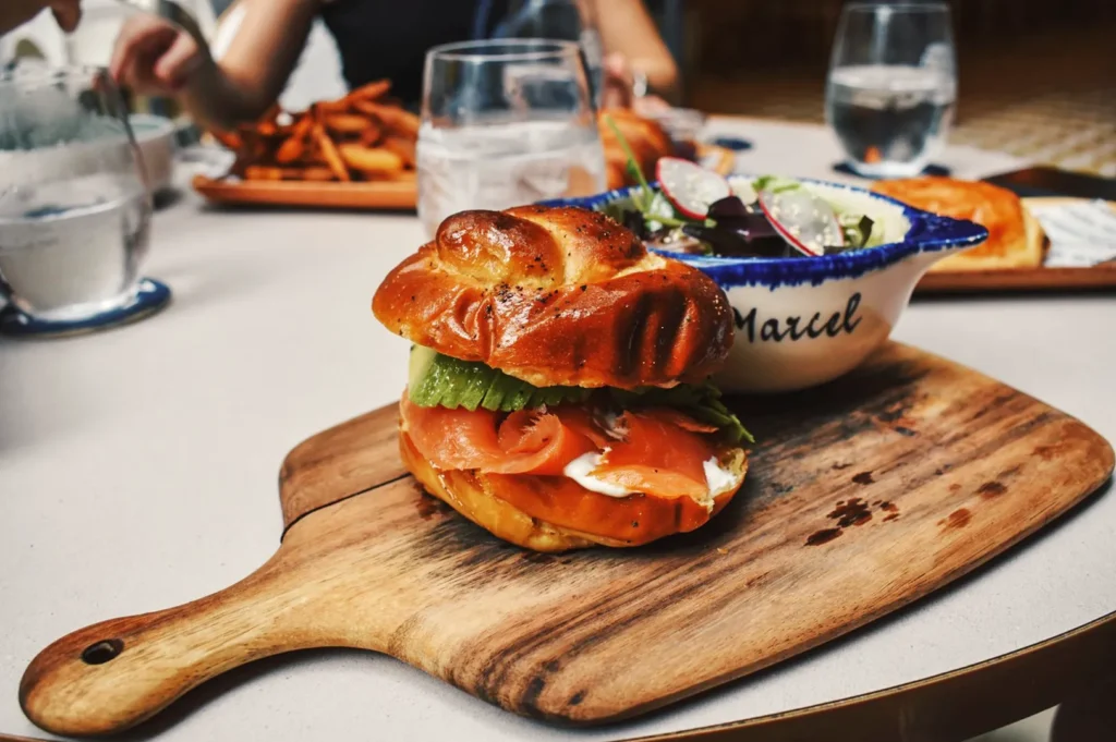 Gourmet smoked salmon sandwich with avocado and cream cheese on a wooden board, served with fresh salad at Merci Marcel café in Singapore.