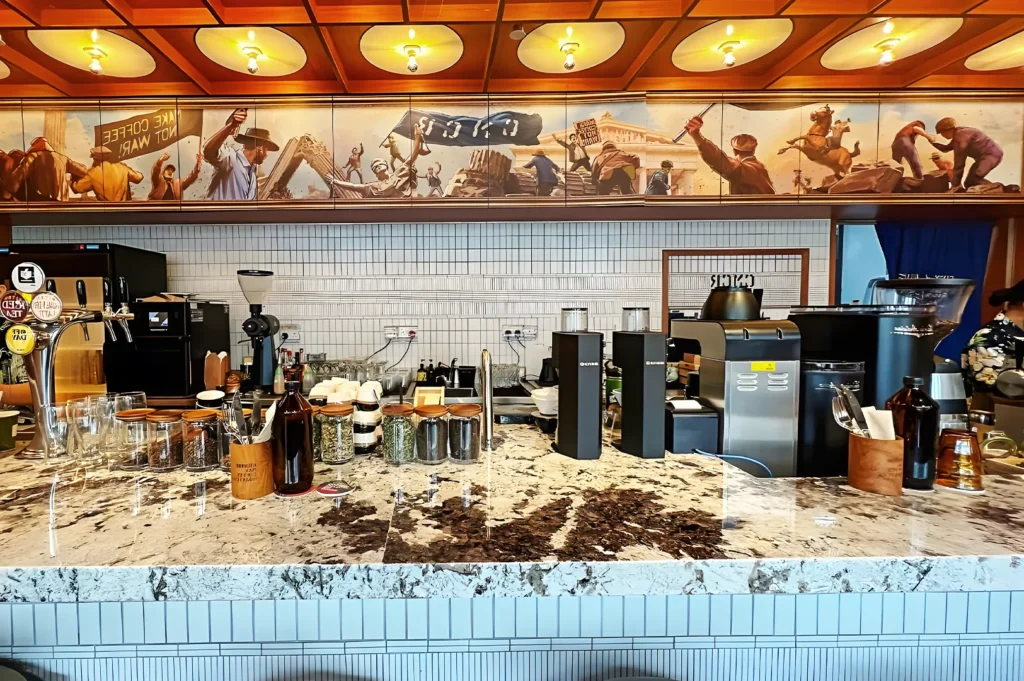 Artistic coffee shop counter with marble surface, brewing equipment, and mural reading “Make Coffee Not War” — a creative café interior with modern design.