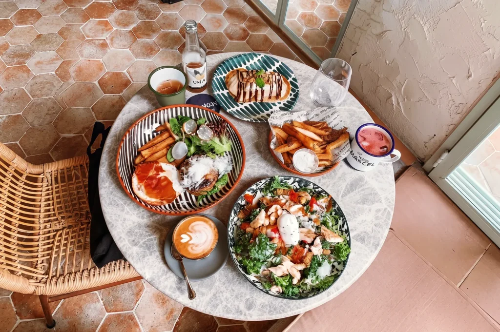 Brunch spread at Merci Marcel café featuring salad with poached egg, burger with fries, dessert waffle, and latte art coffee on a marble table.