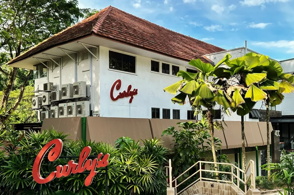 Curly’s restaurant in Singapore surrounded by tropical greenery and palm trees, featuring a red‑tiled roof and bold red signage — modern dining spot with lush outdoor ambiance.