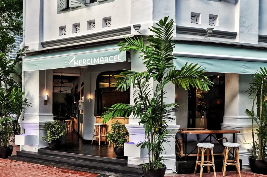 Exterior of Merci Marcel café in Singapore with tropical plants, colonial architecture, and inviting outdoor seating under a pastel green awning.