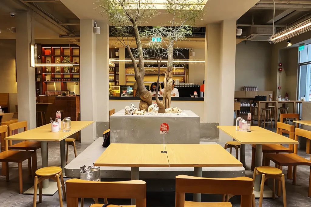 Minimalist café interior in Singapore with wooden furniture, central planter with sculptural trees, and warm lighting — modern dining design blending nature and urban aesthetics.
