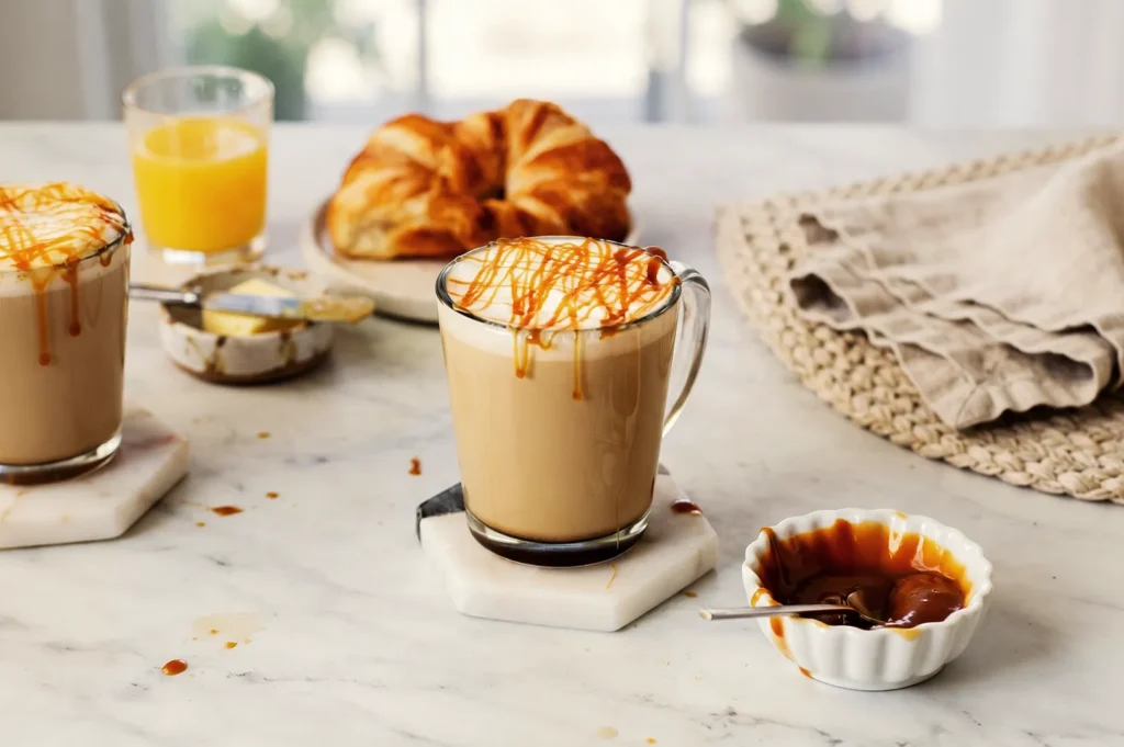 Glass mug filled with a caramel macchiato topped with whipped cream and caramel drizzle on a marble kitchen counter.