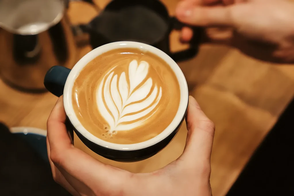 Hands holding a blue ceramic mug filled with a freshly poured leaf latte art design.