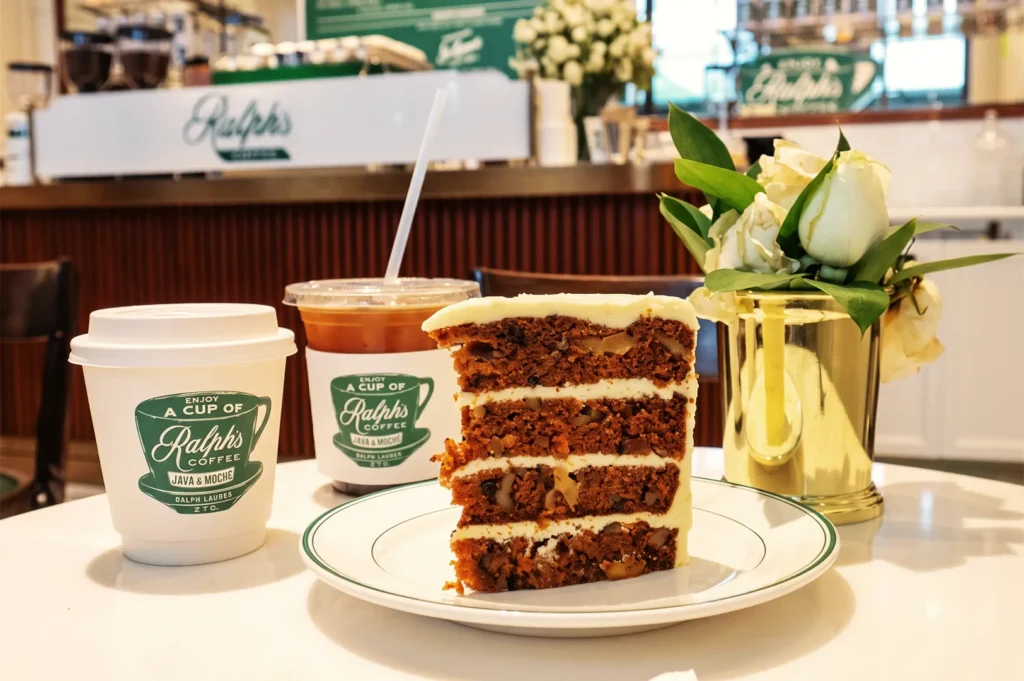 Ralph’s Coffee Singapore serving carrot cake slice with iced and hot coffee in branded cups beside white roses in gold vase.