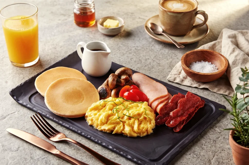 Beanstro Singapore breakfast platter with scrambled eggs, pancakes, bacon, ham, mushrooms, and coffee served with orange juice.