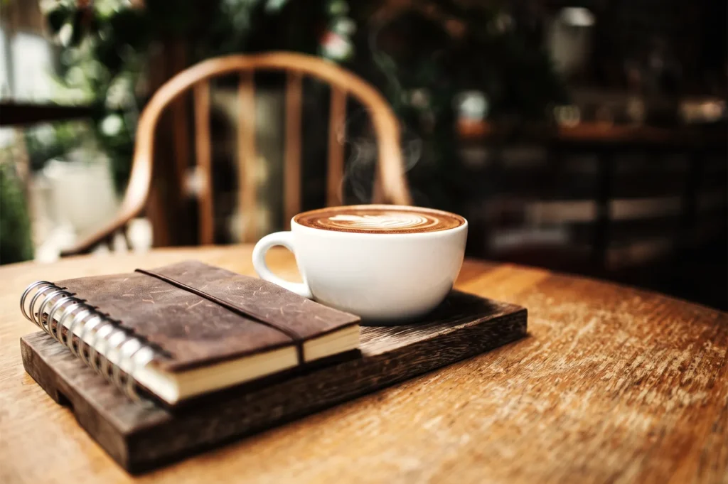 A hot, steaming cup of latte with latte art resting next to a brown leather-bound spiral journal on a wooden tray at a cafe table.