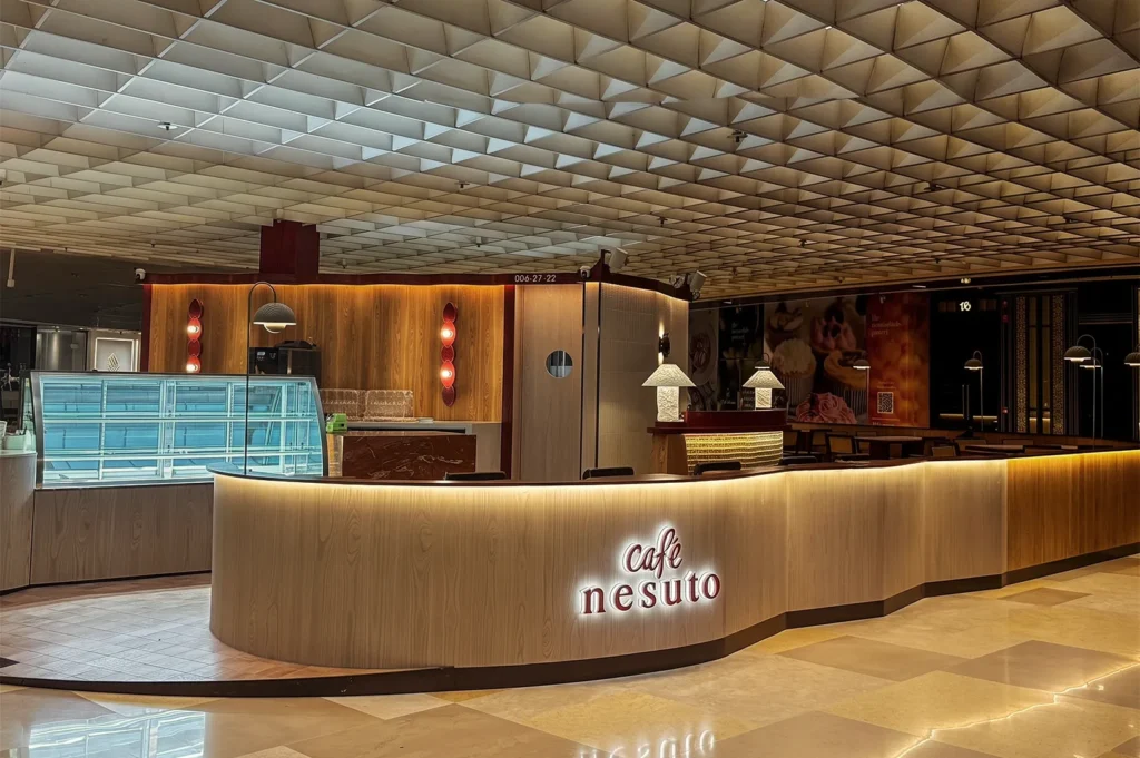 Elegant Café Nesuto Singapore with curved wooden counter, illuminated signage, and modern dessert display under geometric ceiling panels.
