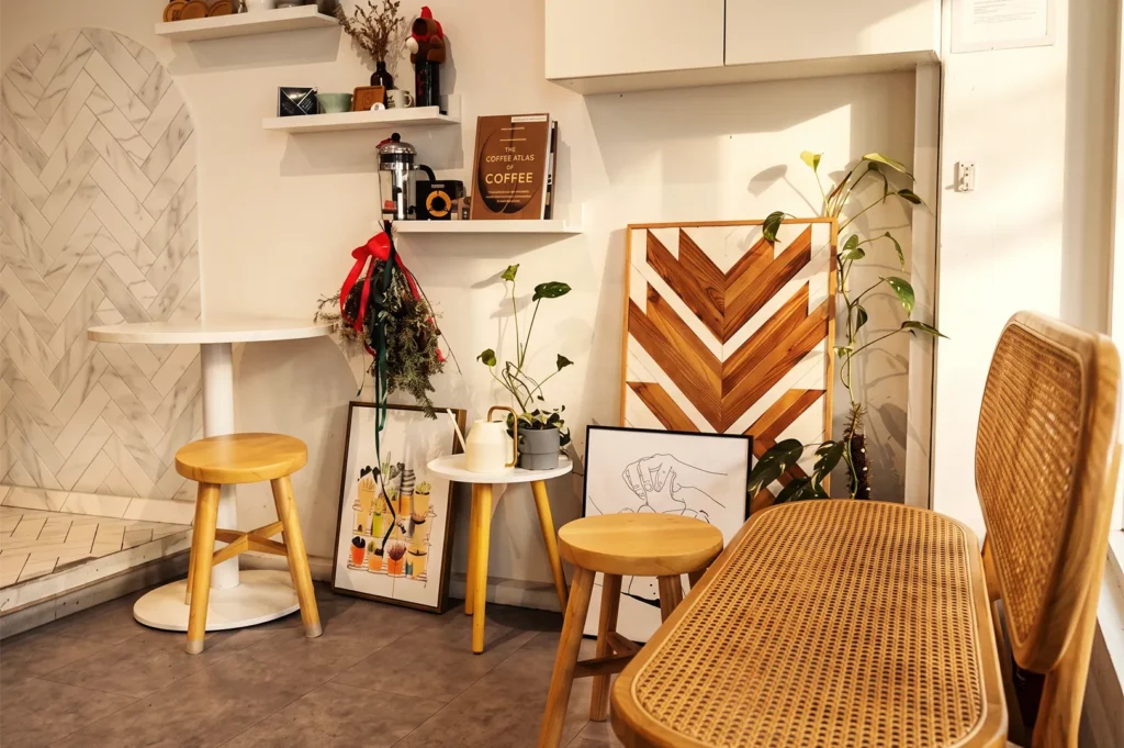 Cafe seating corner decorated with a rattan bench, wooden stools, framed artwork, potted houseplants, and shelving displaying "The World Atlas of Coffee" book.