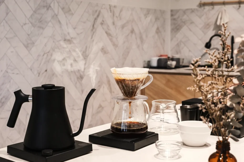 Close-up of artisanal pour-over filter coffee dripping from a V60 dripper into a glass server beside a matte black electric kettle on a digital scale.