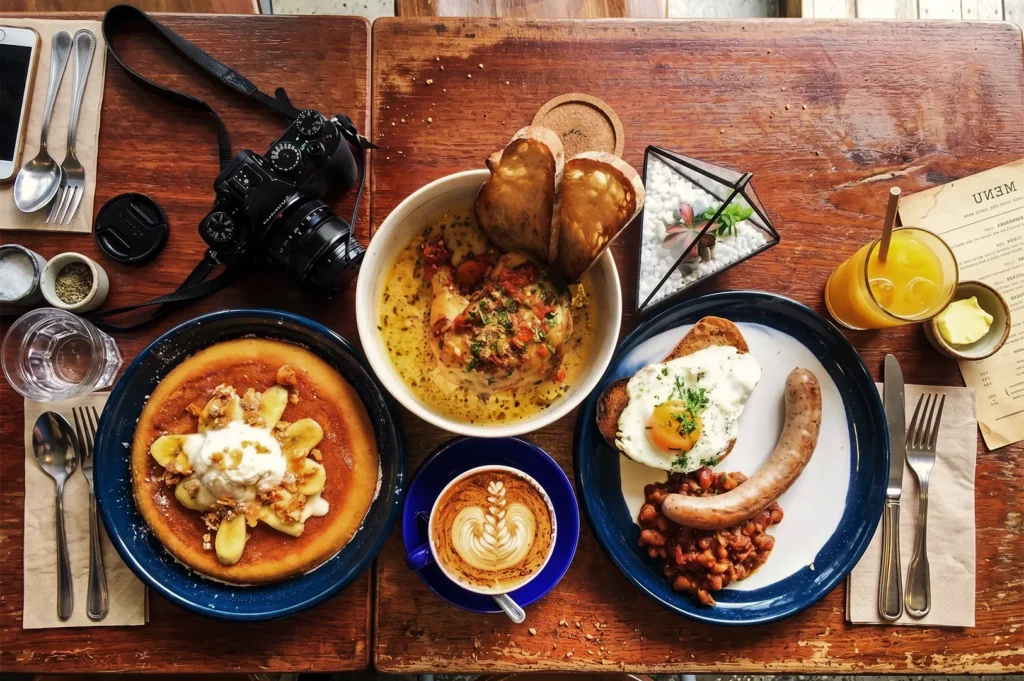 Atlas Coffeehouse Singapore brunch spread with pancakes, eggs, sausage, creamy chicken dish, latte art coffee, and orange juice on wooden table.