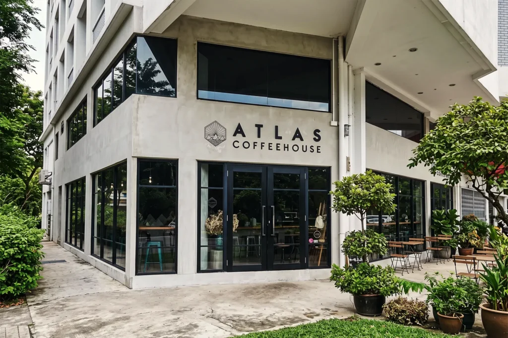 Corner exterior of Atlas Coffeehouse Singapore surrounded by greenery, outdoor seating, and modern concrete architecture with large glass windows.